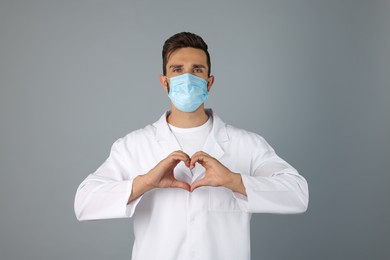 Doctor in protective mask making heart with hands on grey background