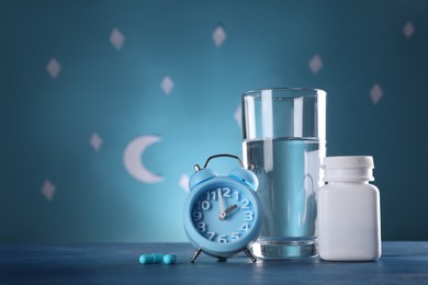 Alarm clock and soporific pills near glass of water on table against blue wall decorated with stars and crescent, space for text. Insomnia treatment