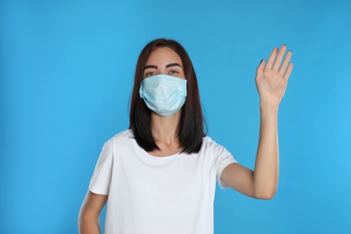 Young woman in protective mask showing hello gesture on light blue background. Keeping social distance during coronavirus pandemic