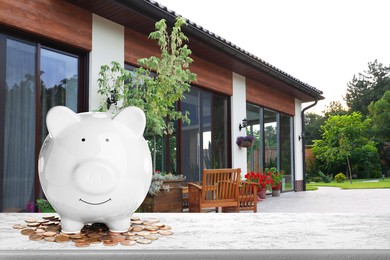 Piggy bank and coins on stone surface and beautiful view of house, space for text. Mortgage concept