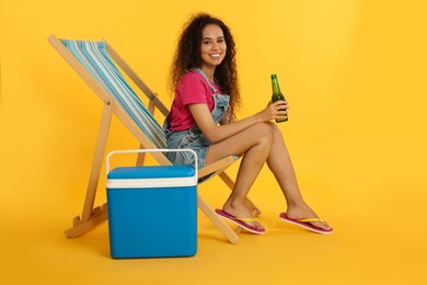 Happy young African American woman with bottle of beer resting in deck chair near cool box on yellow background