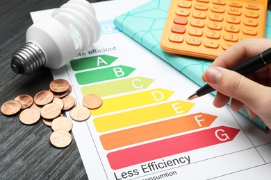 Woman with pen, energy efficiency rating chart, coins, light bulb and calculator at table, closeup