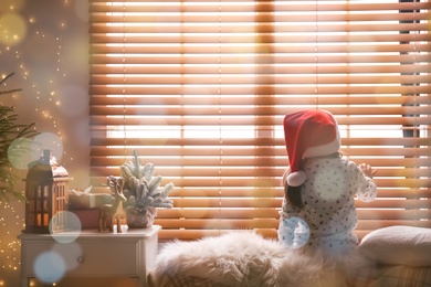 Photo of Little girl in Christmas hat near window at home, back view