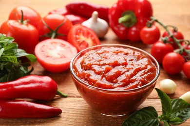 Delicious adjika sauce in glass bowl and ingredients on wooden table, closeup