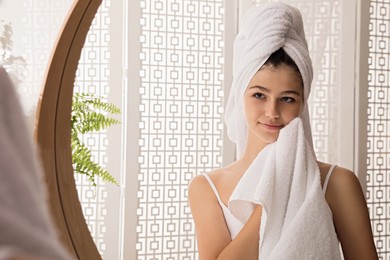 Beautiful teenage girl wiping face with towel near mirror at home