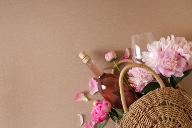 Wicker bag with bottle of rose wine and beautiful pink peonies on brown background, top view. Space for text