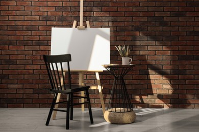 Photo of Wooden easel with blank canvas and different art supplies near brick wall in room