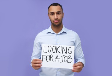 Photo of Young unemployed man holding sign with phrase Looking For A Job on violet background