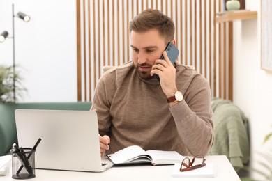 Photo of Young man talking on phone while working with laptop at home