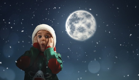 Surprised little girl in Santa hat on full moon night. Christmas holiday 