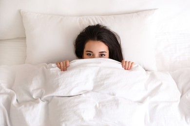 Young woman hiding under warm white blanket in bed, top view