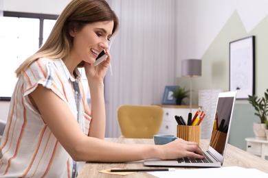 Young woman talking on phone while working with laptop at desk in home office