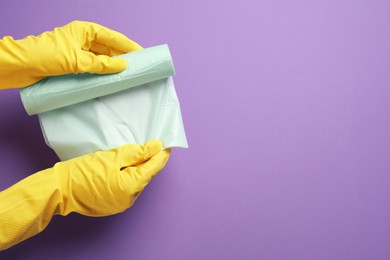Janitor in rubber gloves holding roll of color garbage bags over violet background, top view. Space for text