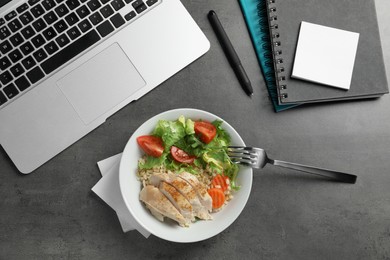 Photo of Bowl with tasty food, laptop, fork and notebooks on grey table, flat lay. Business lunch