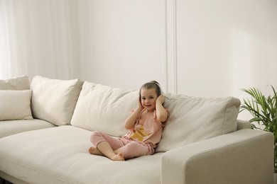 Little girl with headphones sitting on comfortable sofa in living room