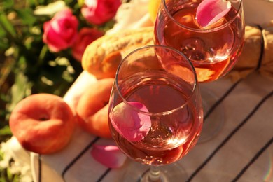 Photo of Glasses of delicious rose wine with petals, baguette and peaches on white picnic blanket outside, closeup