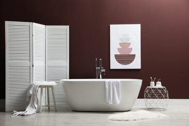 Modern ceramic bathtub and furniture near burgundy wall in room