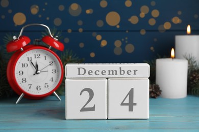 Christmas Eve - December 24. Block calendar, alarm clock and burning candles on light blue wooden table