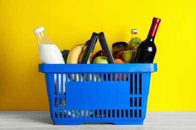 Shopping basket with grocery products on white wooden table against yellow background