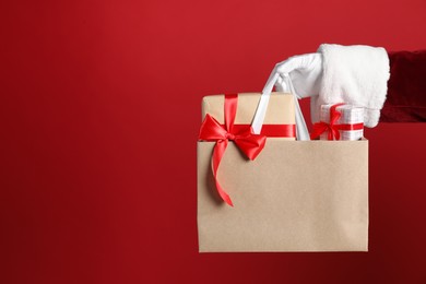 Photo of Santa holding paper bag with gift boxes on red background, closeup. Space for text