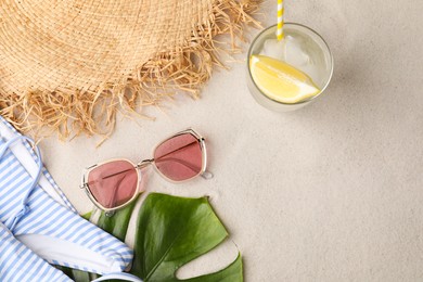 Stylish sunglasses, glass of refreshing drink and beach accessories on sand, flat lay