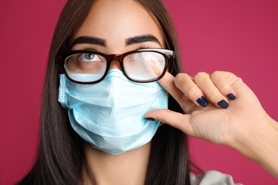 Young woman wiping foggy glasses caused by wearing disposable mask on pink background. Protective measure during coronavirus pandemic