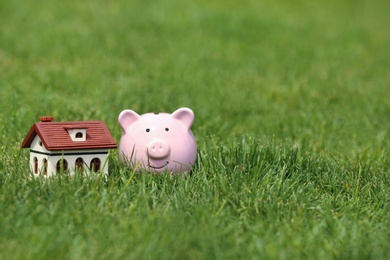 Piggy bank and model of house on green grass outdoors. Space for text