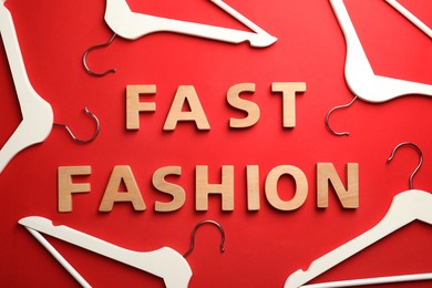 Phrase FAST FASHION made of wooden letters and white hangers on red background, flat lay