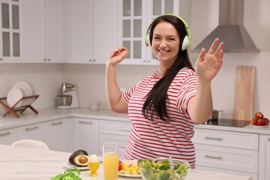 Happy overweight woman with headphones dancing near table in kitchen. Healthy diet