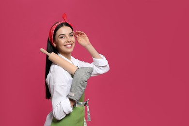 Photo of Young housewife in oven glove holding rolling pin on pink background. Space for text