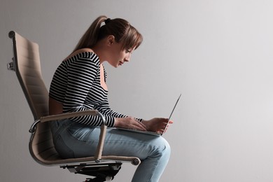 Young woman with poor posture using laptop while sitting on chair against grey background, space for text