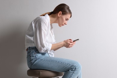 Woman with bad posture using smartphone while sitting on stool against light grey background