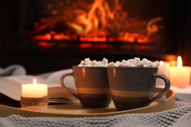 Cups of hot drink, candle and open book near fireplace indoors