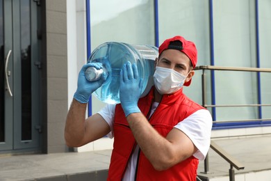 Courier in medical mask holding bottle for water cooler near building outdoors. Delivery during coronavirus quarantine