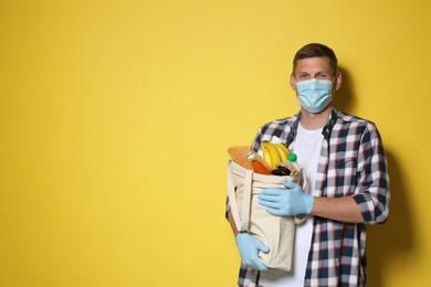 Male volunteer in protective mask and gloves with products on yellow background, space for text. Aid during coronavirus quarantine