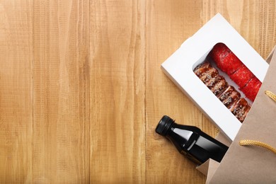 Photo of Box with delicious sushi rolls and bottle of soy sauce in paper package on wooden table, flat lay. Space for text