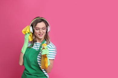 Beautiful young woman with headphones, rag and bottle of detergent singing on pink background. Space for text