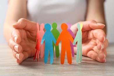 Photo of Woman protecting paper human figures on wooden table, closeup. Diversity and inclusion concept