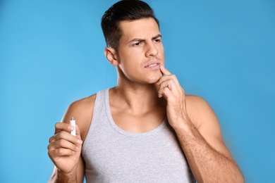 Photo of Emotional man with herpes applying cream on lips against light blue background
