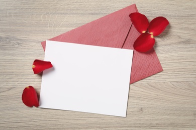Blank greeting card, envelope and rose petals on wooden table, flat lay. Valentine's day celebration