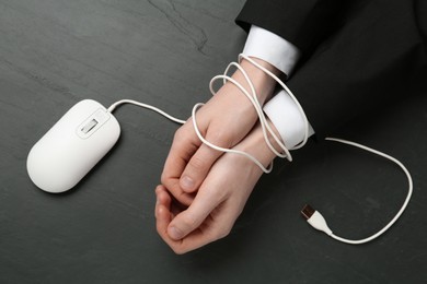 Man showing hands tied with computer mouse cable at black table, top view. Internet addiction