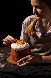 Young woman decorating traditional Easter cake at table against black background