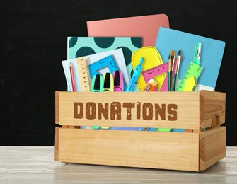 Donation box with different school stationery on white wooden table near blackboard
