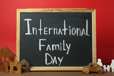 Photo of Small chalkboard with text International Family Day, wooden house models, people figure and toy car on brown table against red background