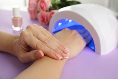 Woman using ultraviolet lamp to dry gel nail polish at violet table, closeup