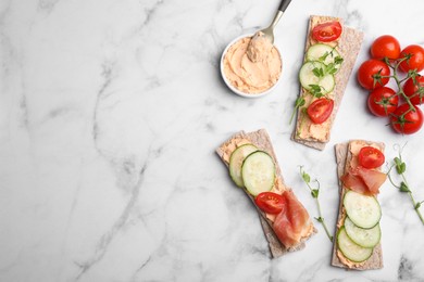 Tasty crispbreads with prosciutto, cream cheese and vegetables on white marble table, flat lay. Space for text