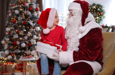 Santa Claus giving present to little girl in room decorated for Christmas
