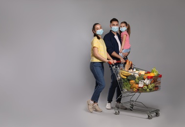 Family with protective masks and shopping cart full of groceries on light grey background