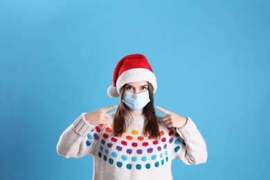 Pretty woman in Santa hat and medical mask on light blue background