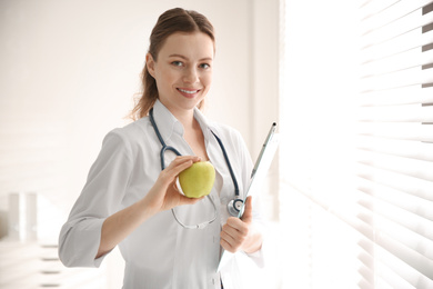 Nutritionist with apple and clipboard near window in office. Space for text